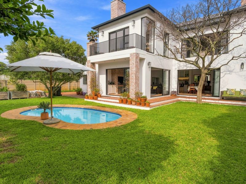 Rear view of a two-story house featuring a landscaped garden, swimming pool, brick patio, and an upper-level balcony with black railings.