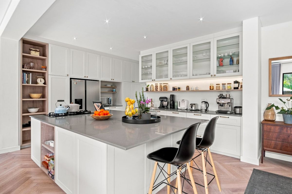 Modern kitchen featuring a large central island with stone countertops, shaker-style cabinetry, glass-front display cabinets, and elegant herringbone timber flooring.