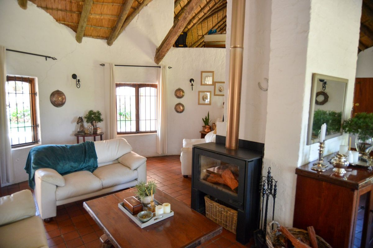 A cozy living room featuring a traditional thatched roof with exposed timber beams, terracotta flooring, and a prominent freestanding fireplace.
