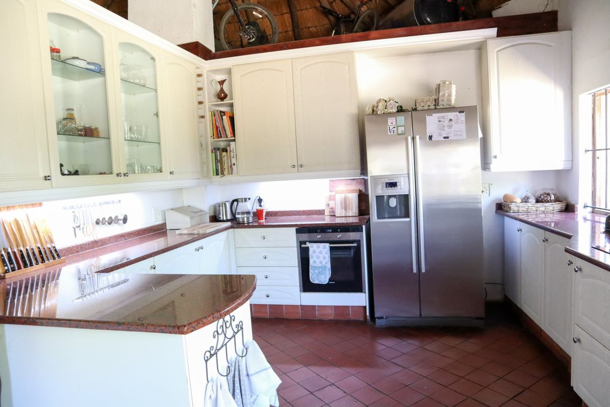 A functional kitchen featuring cream-colored cabinetry, granite countertops, built-in oven, and traditional terracotta tile flooring under a high thatched roof.