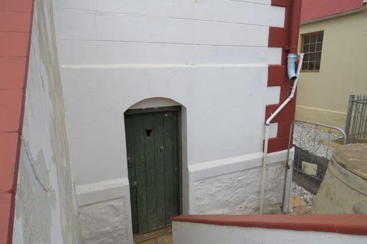 A rear exterior view showing a stone foundation, painted masonry wall, and a weathered timber door with a downpipe.
