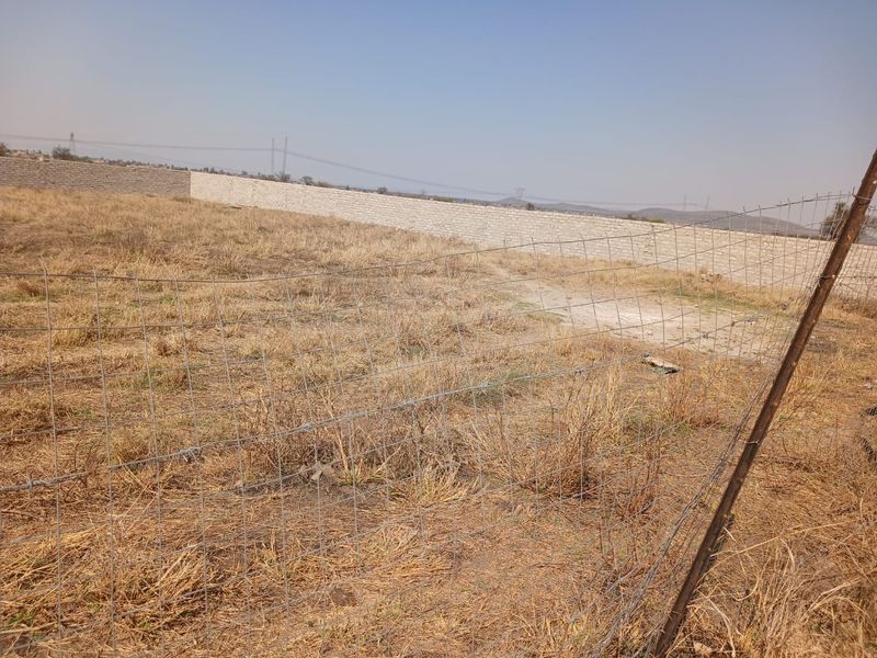 A large, undeveloped plot of land enclosed by wire mesh fencing and a perimeter masonry wall, featuring dry, overgrown vegetation.