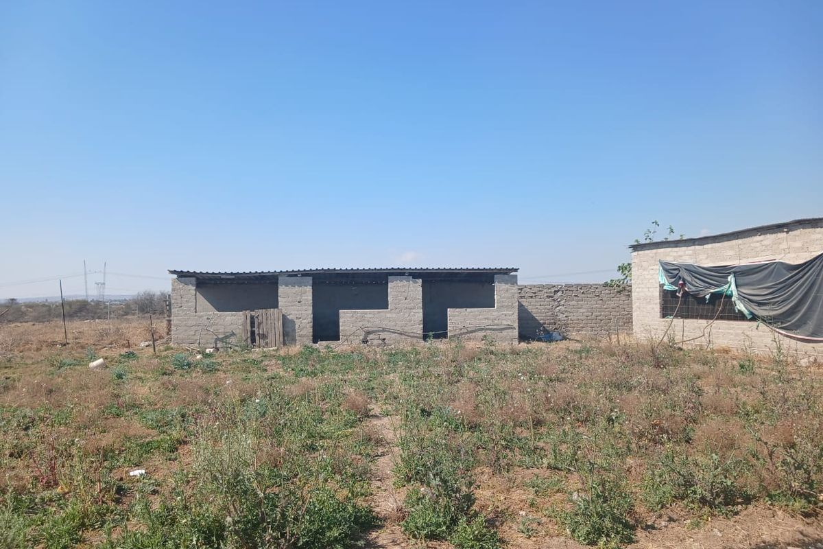 An exterior view of an unfinished concrete block structure on a rural farm property, featuring a corrugated metal roof and surrounding land.