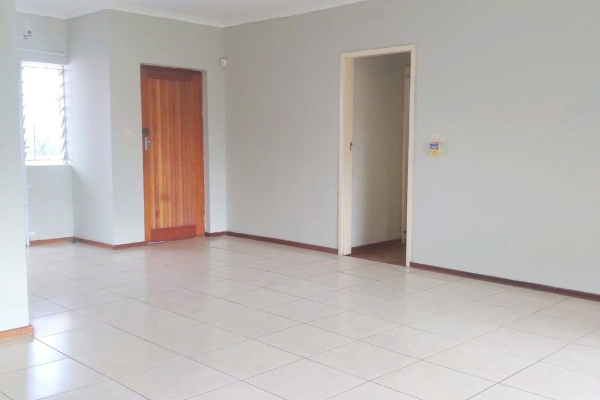 A spacious, empty living area featuring neutral tiled flooring, a ceiling fan, and access to adjacent rooms through wooden and open doorways.