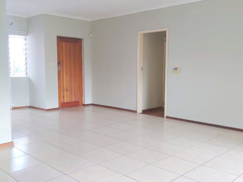 A spacious, empty living area featuring neutral tiled flooring, a ceiling fan, and access to adjacent rooms through wooden and open doorways.