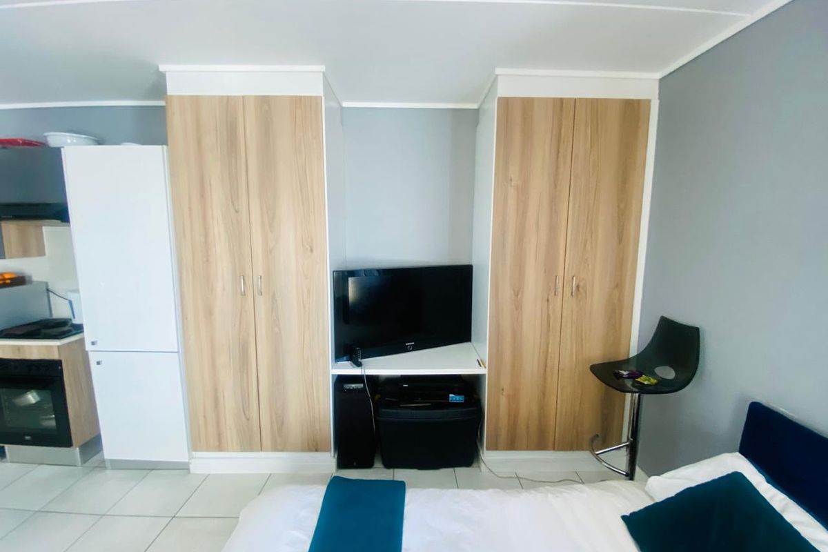 A compact bedroom featuring built-in wood-finish cupboards and tiled flooring, with an integrated kitchen area visible in the background.