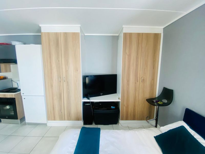 A compact bedroom featuring built-in wood-finish cupboards and tiled flooring, with an integrated kitchen area visible in the background.