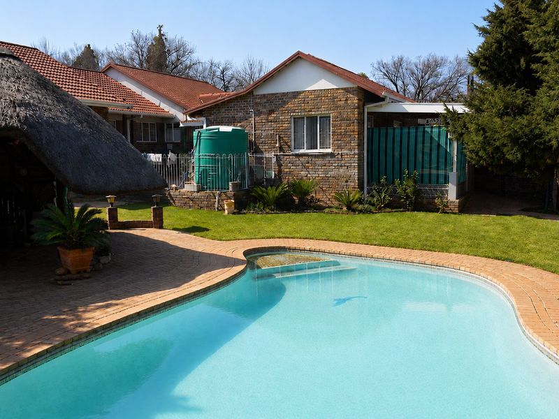 A well-maintained backyard featuring a swimming pool, a thatched lapa, and a brick-faced house with a water storage tank.