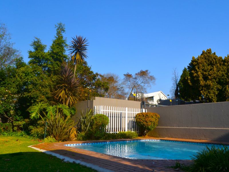 A well-maintained outdoor pool area featuring a brick-edged swimming pool, surrounded by a landscaped garden and secure perimeter walling.