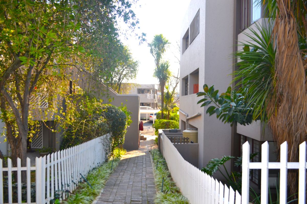 A paved walkway leads between apartment building blocks, featuring white picket fencing and established garden landscaping.