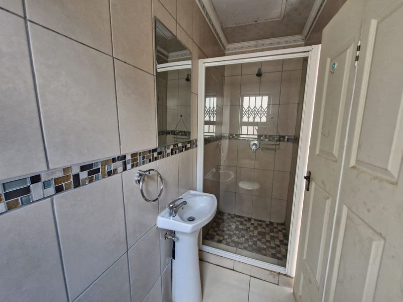 A functional bathroom featuring a pedestal basin, a glass-enclosed shower, and neutral wall tiling with a decorative mosaic border.