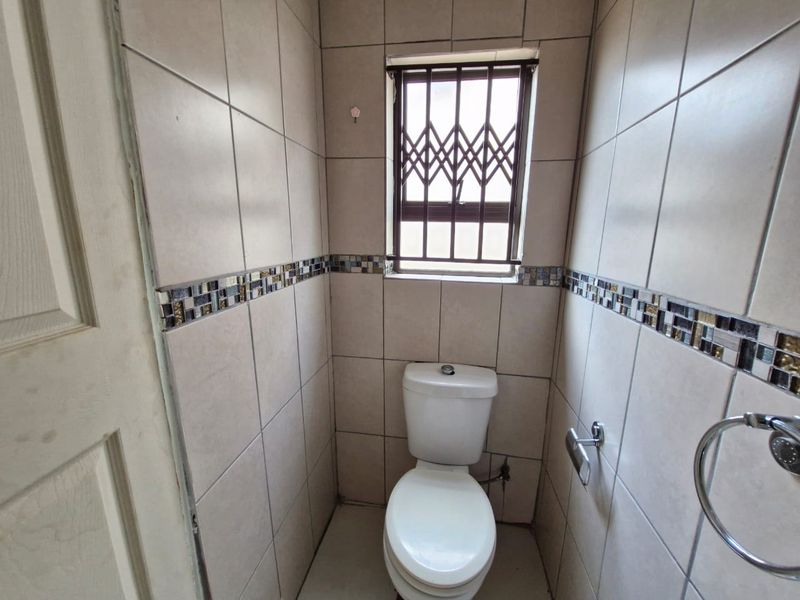 A compact, fully tiled guest bathroom featuring a standard toilet, mosaic accent border, and a window fitted with a security gate.