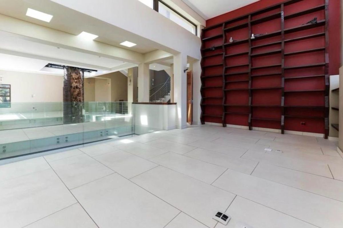 Spacious double-volume living area featuring large-format floor tiles, a striking floor-to-ceiling built-in shelving unit, and a modern glass balustrade overlooking the lower level.