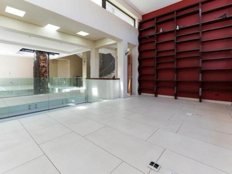 Spacious double-volume living area featuring large-format floor tiles, a striking floor-to-ceiling built-in shelving unit, and a modern glass balustrade overlooking the lower level.