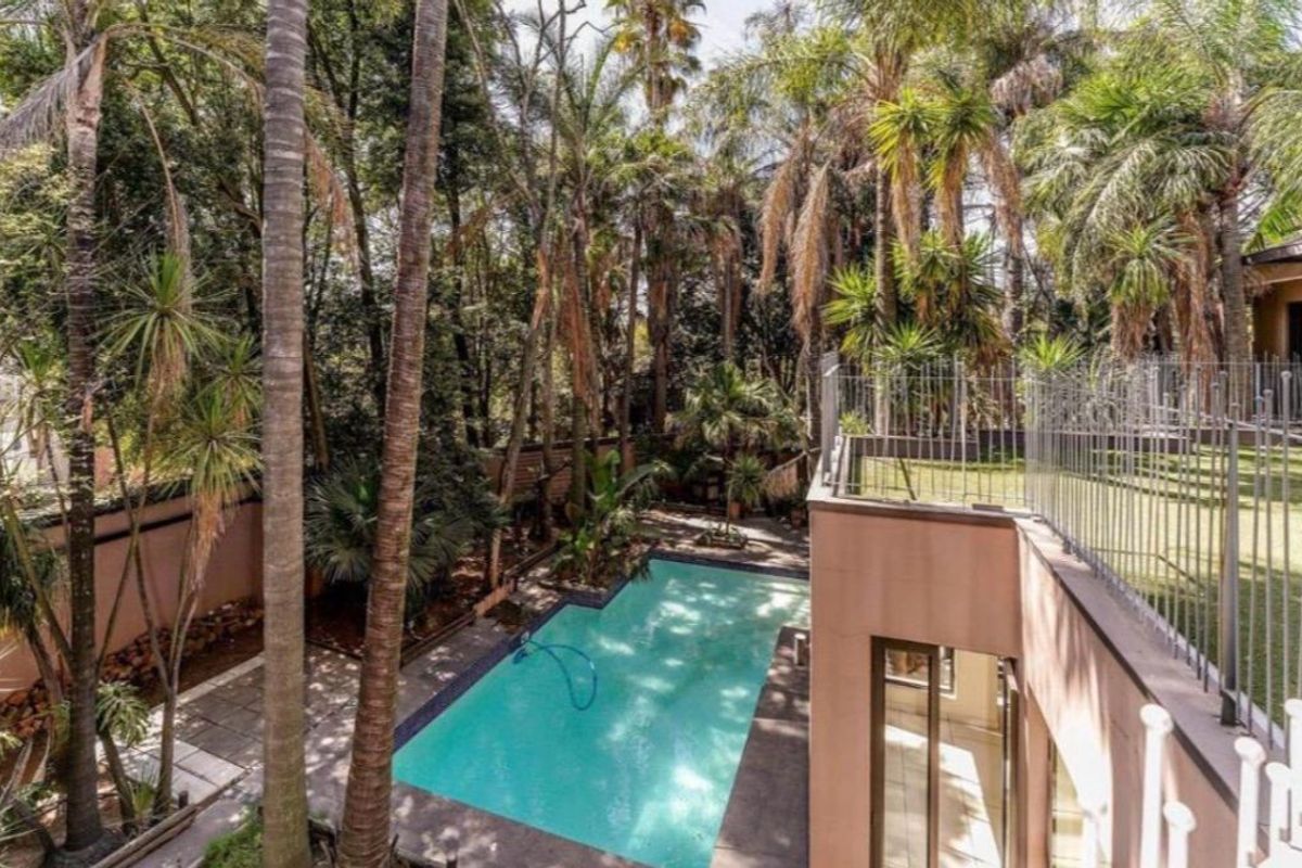 Outdoor pool area featuring a swimming pool, paved surround, and metal perimeter fencing, set within a mature, tree-lined garden environment.