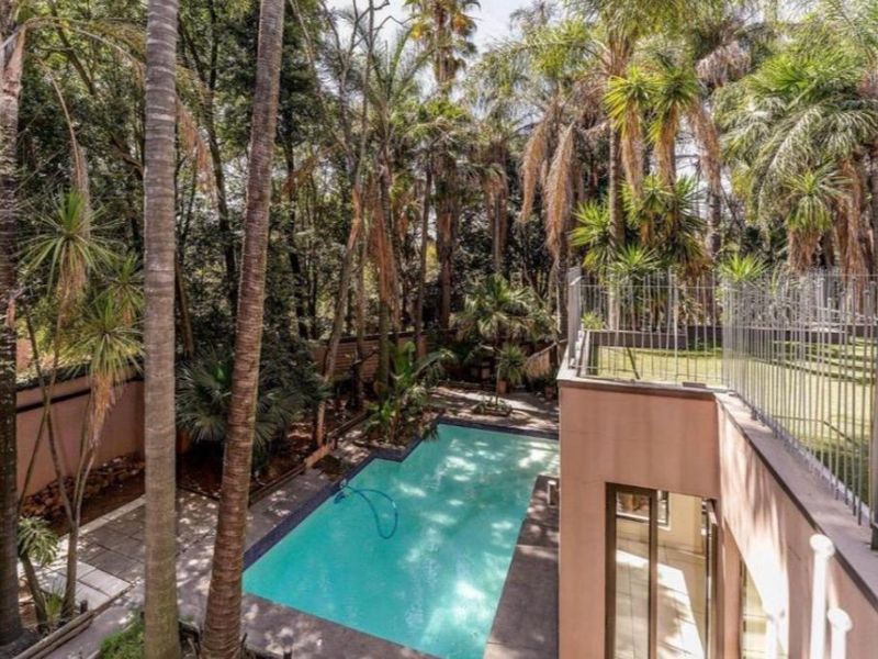Outdoor pool area featuring a swimming pool, paved surround, and metal perimeter fencing, set within a mature, tree-lined garden environment.