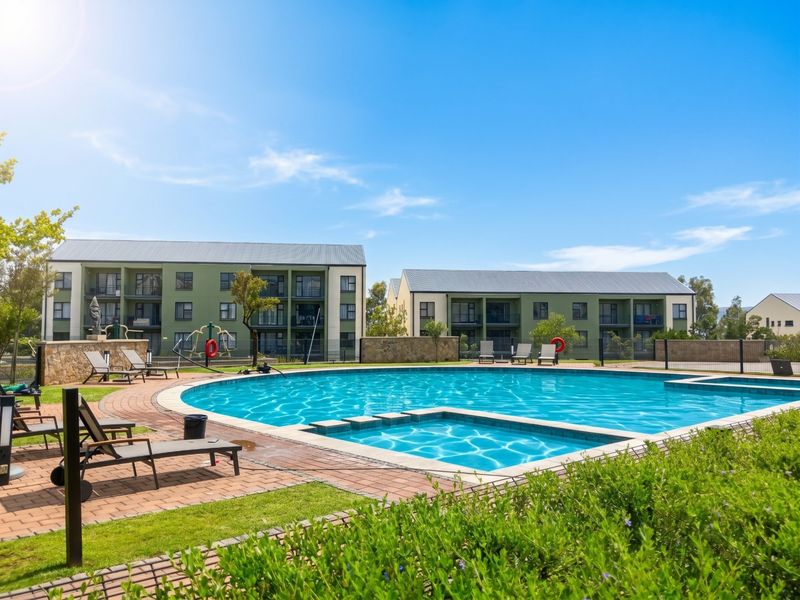 A well-maintained communal swimming pool area featuring paved surrounds, landscaped gardens, and views of the apartment complex buildings.