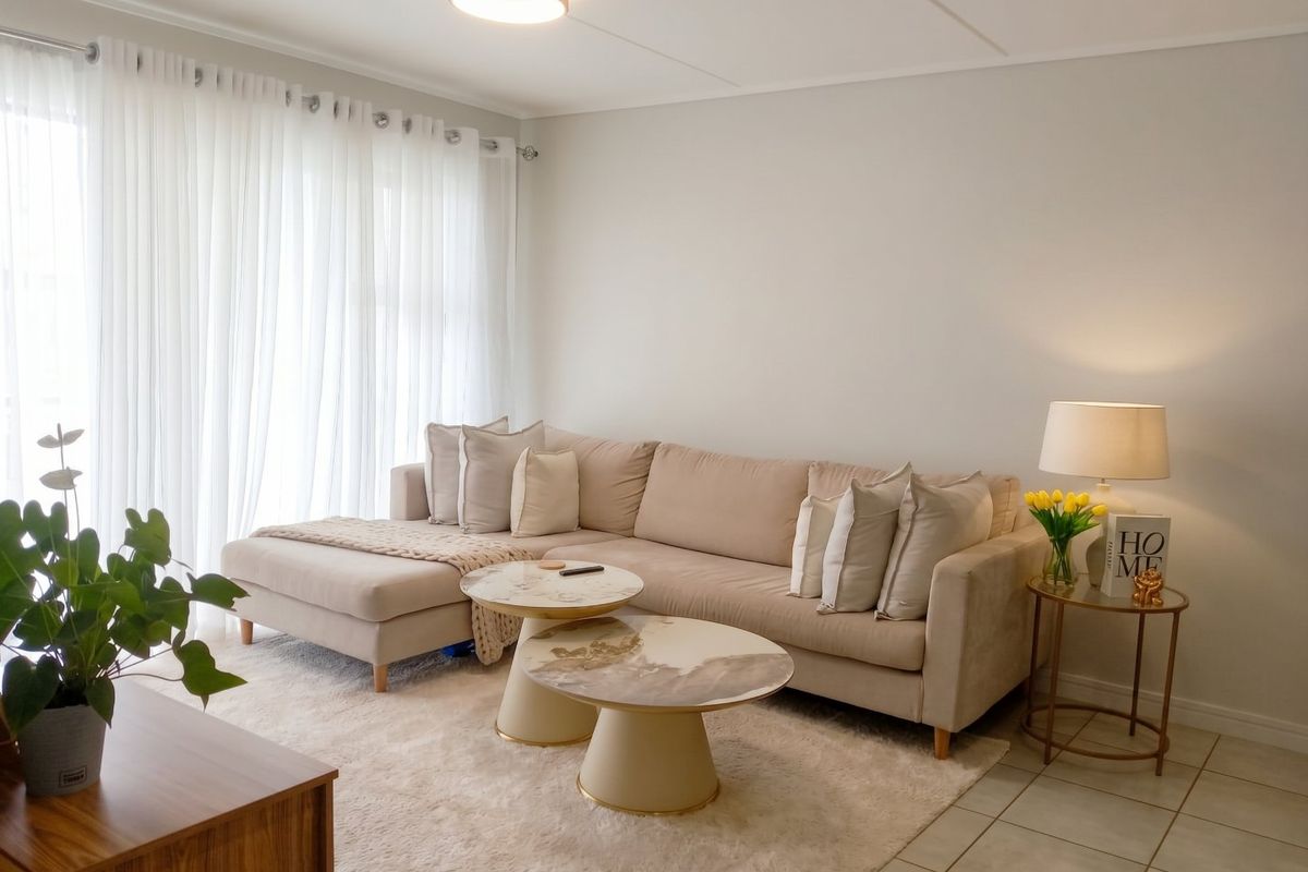 A bright and well-maintained living room featuring neutral tiled flooring, modern ceiling lighting, and large windows dressed with sheer curtains.