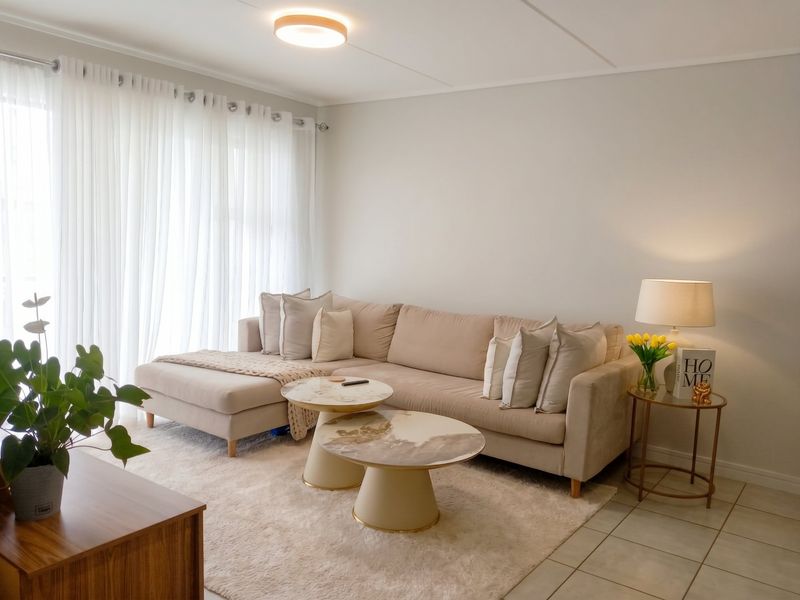 A bright and well-maintained living room featuring neutral tiled flooring, modern ceiling lighting, and large windows dressed with sheer curtains.
