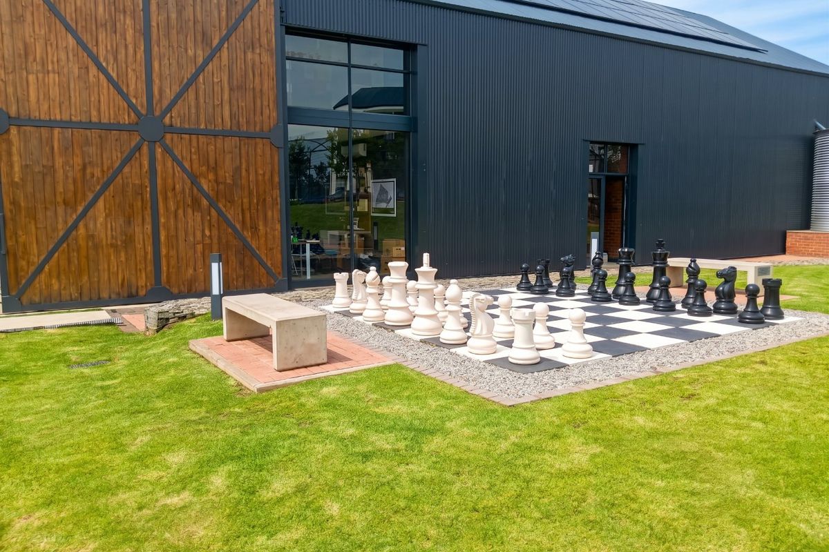 Modern exterior facade featuring corrugated metal cladding, timber accents, and a landscaped outdoor area with a large-scale chess board and concrete seating.