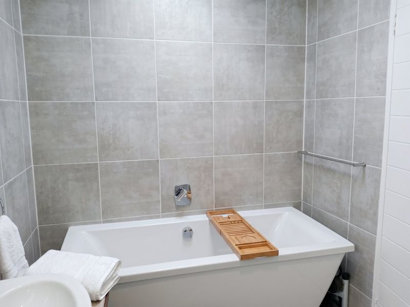 A modern bathroom featuring a clean white built-in bathtub with wall-mounted chrome fixtures and neutral grey ceramic wall tiles.