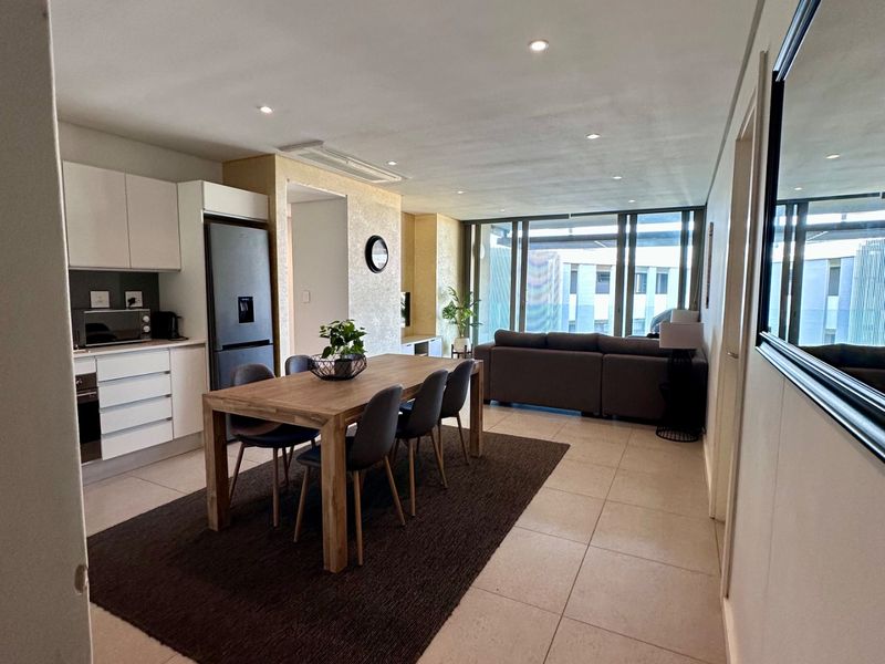 Modern open-plan living and dining area featuring tiled floors, recessed lighting, and a sleek kitchen with integrated appliances leading to a balcony.
