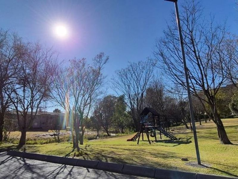 A well-maintained communal garden area featuring a paved walkway, mature trees, and a playground set within an apartment complex.