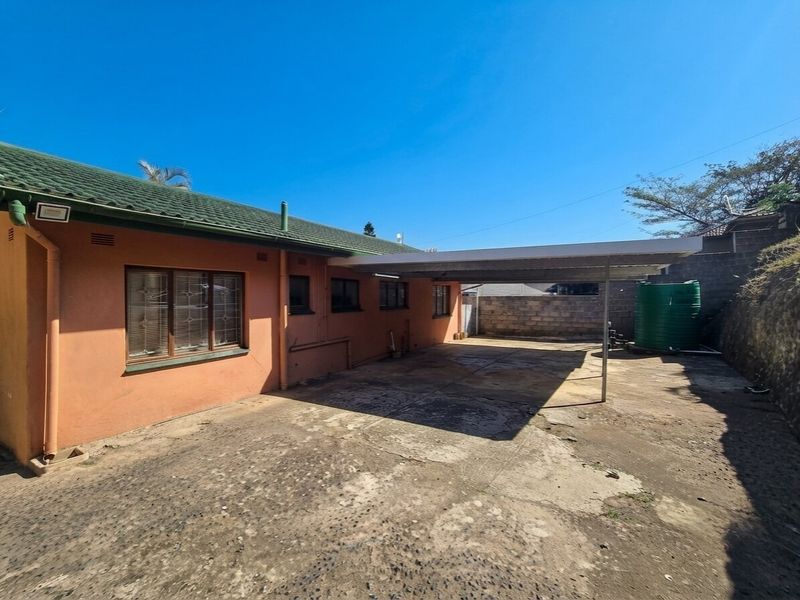 Rear exterior view featuring a concrete driveway, a covered carport, and a water storage tank against a brick boundary wall.