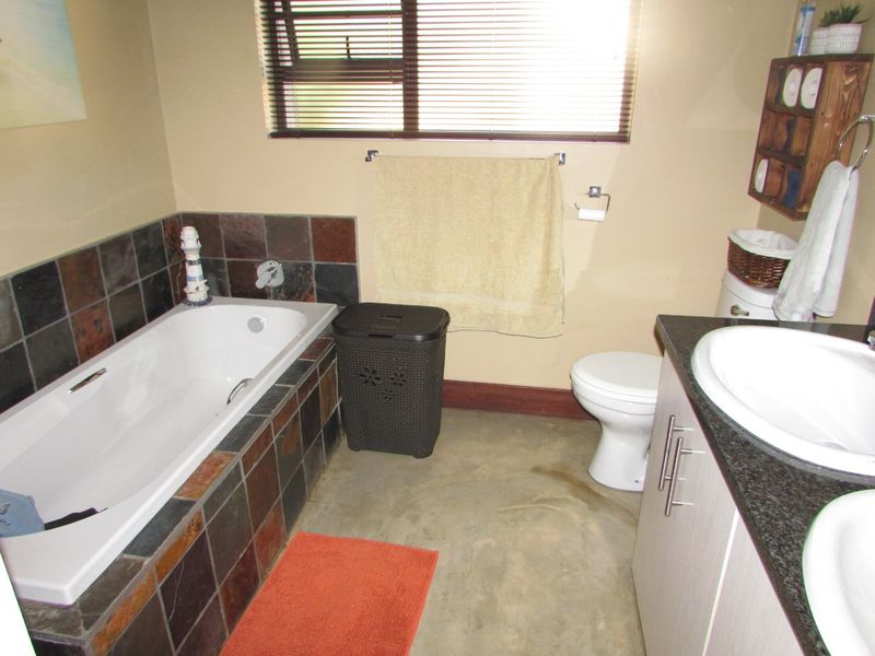 A functional bathroom featuring a built-in bathtub with slate tile surround, a dual vanity with granite countertop, and a toilet.