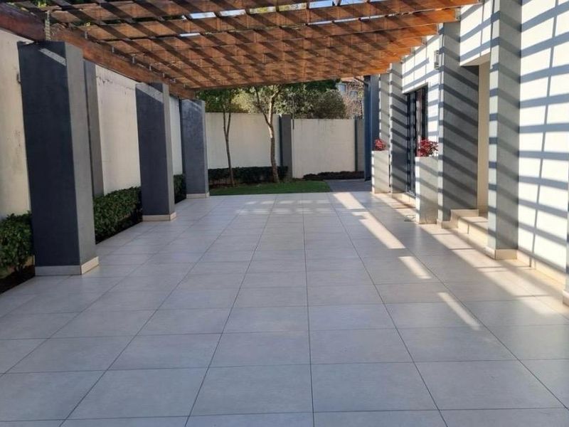 A spacious covered patio featuring a wooden pergola, tiled flooring, and modern masonry pillars, offering a clean and well-maintained outdoor entertainment area.
