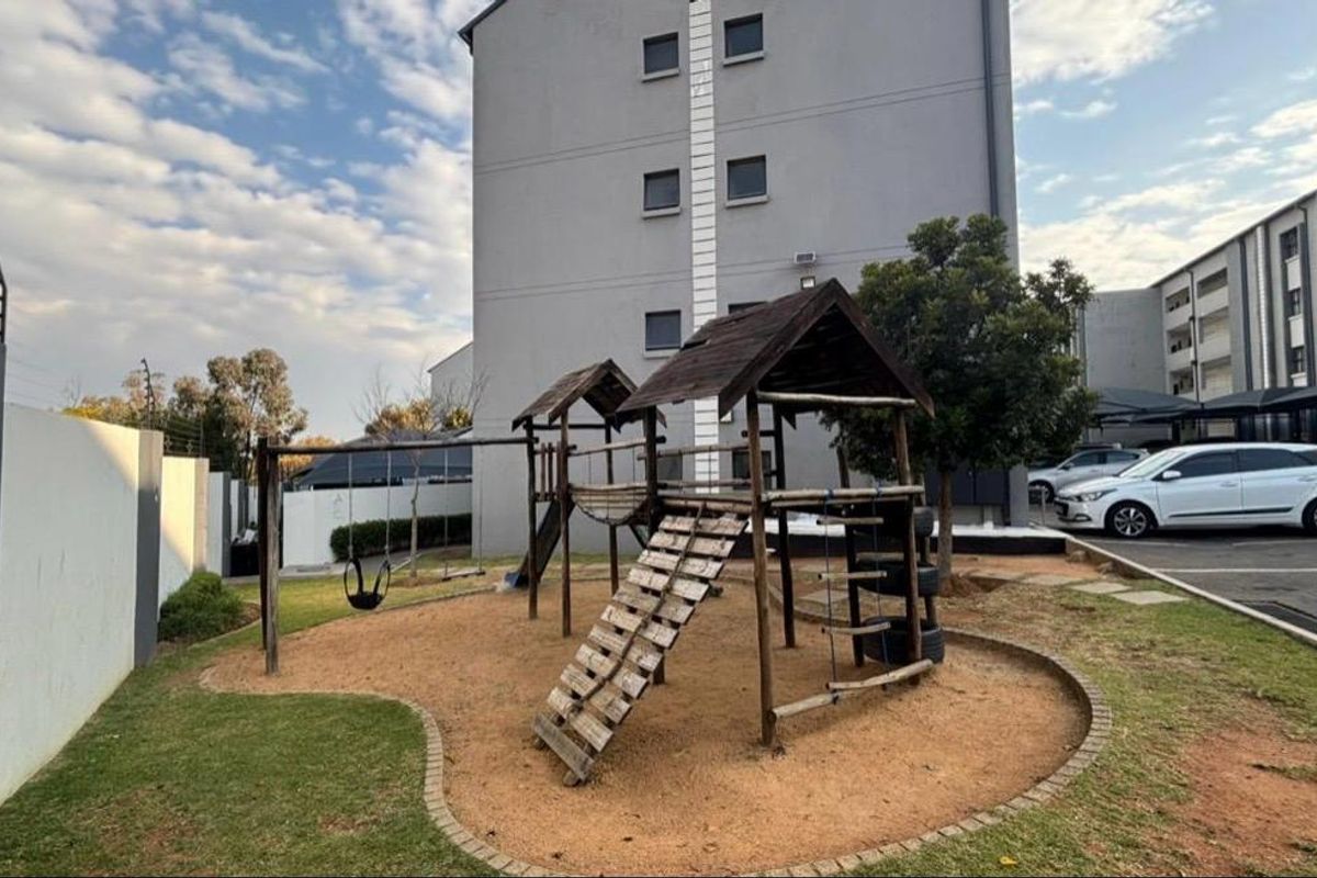 Communal outdoor area featuring a playground with wooden structures, set against a multi-story apartment building with adjacent paved parking.