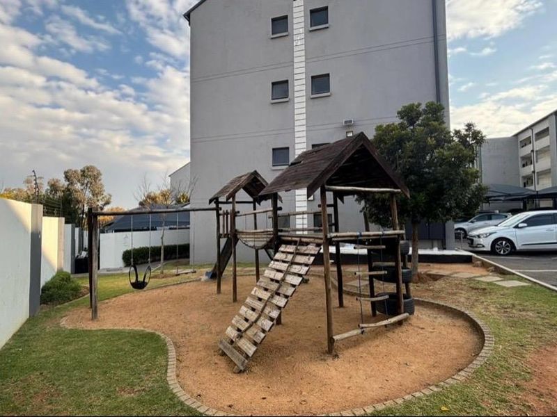 Communal outdoor area featuring a playground with wooden structures, set against a multi-story apartment building with adjacent paved parking.