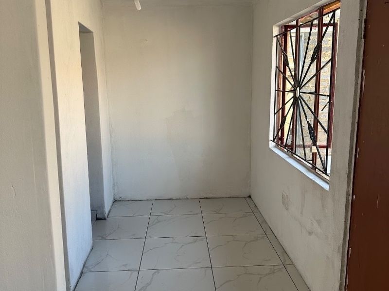 An empty hallway featuring white tiled flooring, a window with security bars, and basic lighting, requiring finishing touches to the walls and door frames.