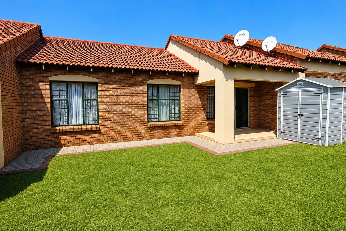 A well-maintained rear exterior view of a face brick townhouse featuring a tiled roof, paved walkway, and a neat lawn area.