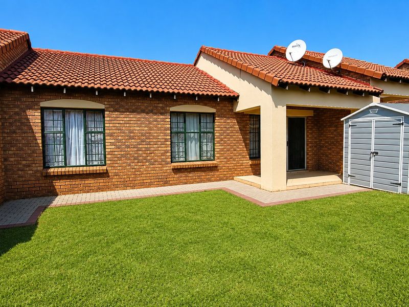 A well-maintained rear exterior view of a face brick townhouse featuring a tiled roof, paved walkway, and a neat lawn area.