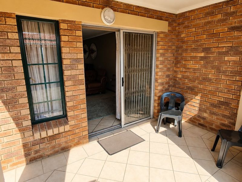 A covered patio area featuring exposed brick walls, tiled flooring, and a sliding glass door with a security gate providing access to the interior.