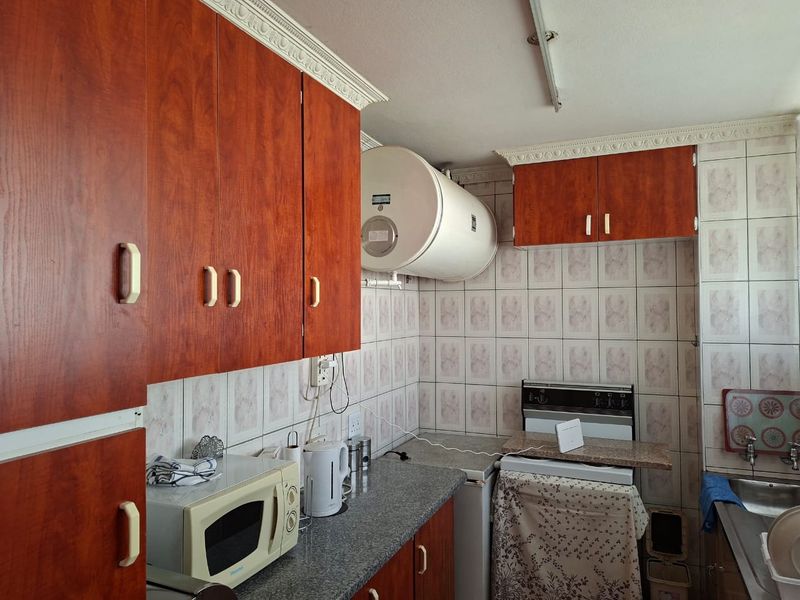 A compact kitchen featuring dated wood-look cabinetry, ceramic wall tiling, a built-in electric geyser, and a functional layout with a stainless steel sink.