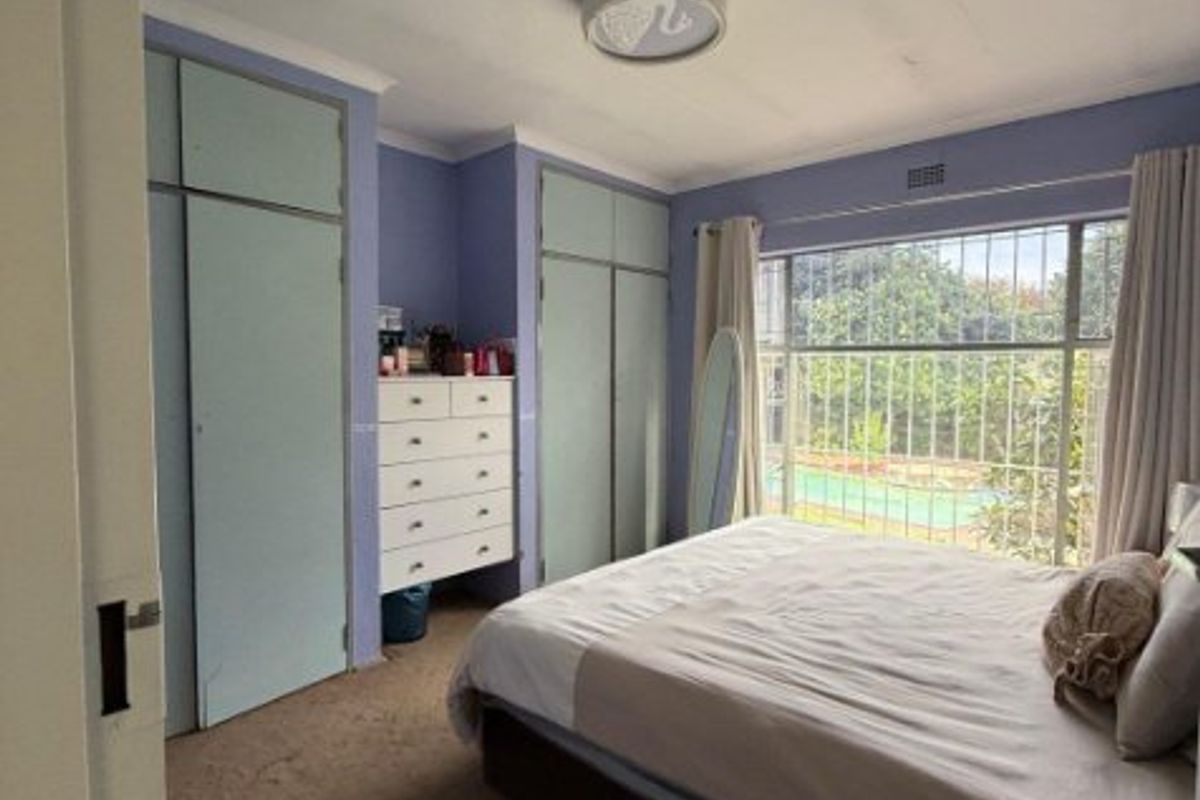 A bedroom featuring built-in cupboards, carpet flooring, and large windows with security bars, offering a functional space with potential for cosmetic updates.