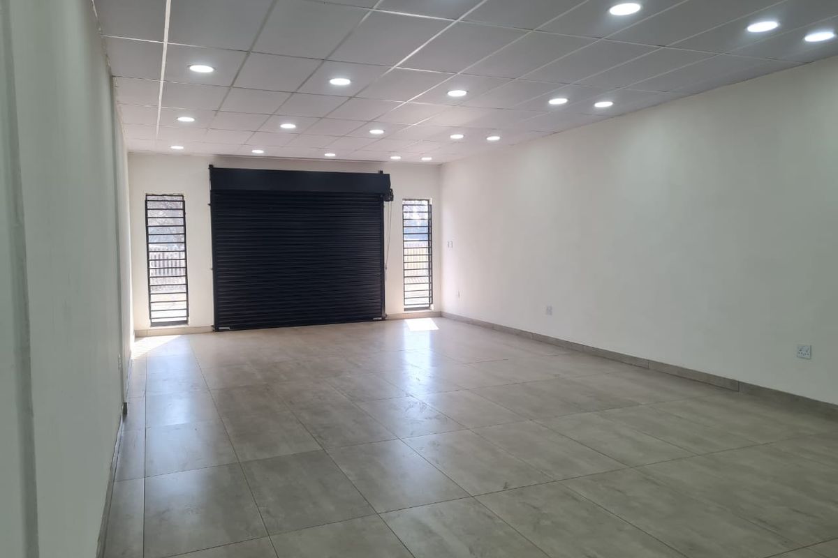 A spacious, empty commercial open-plan unit featuring tiled flooring, a suspended ceiling with recessed lighting, and a secure roller shutter entrance.