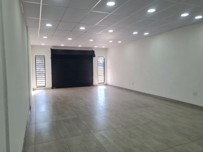 A spacious, empty commercial open-plan unit featuring tiled flooring, a suspended ceiling with recessed lighting, and a secure roller shutter entrance.