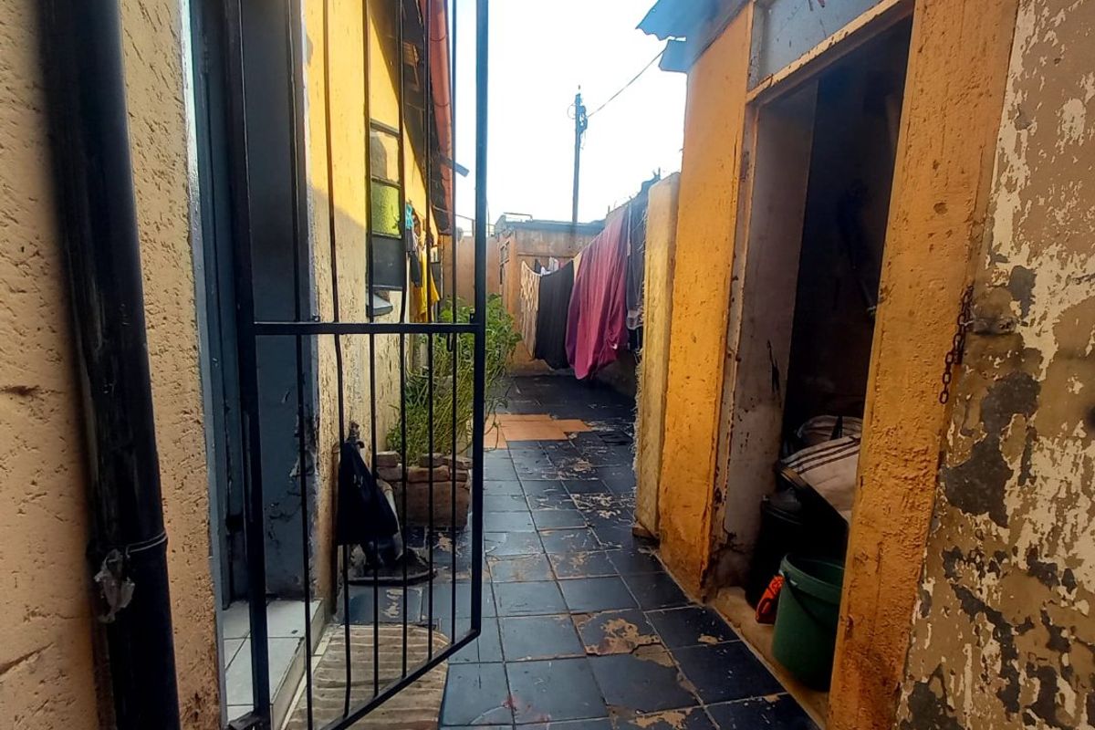 A narrow exterior rear passage featuring tiled flooring and yellow painted walls, showing significant signs of wear and maintenance requirements.