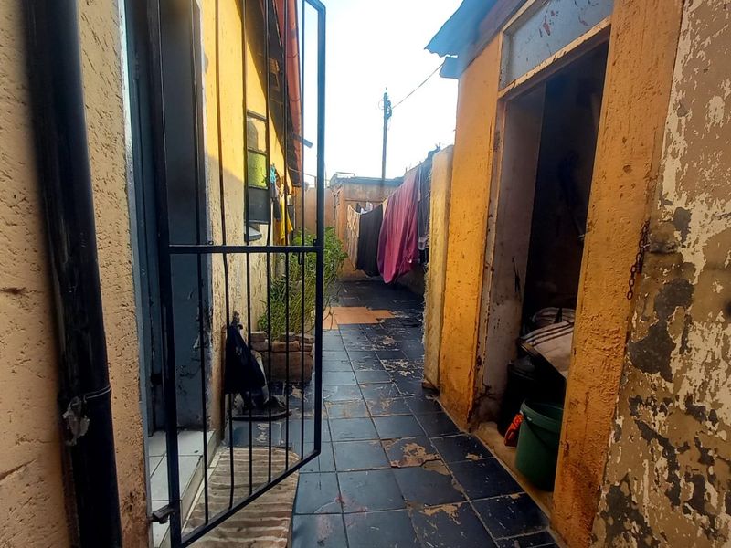 A narrow exterior rear passage featuring tiled flooring and yellow painted walls, showing significant signs of wear and maintenance requirements.
