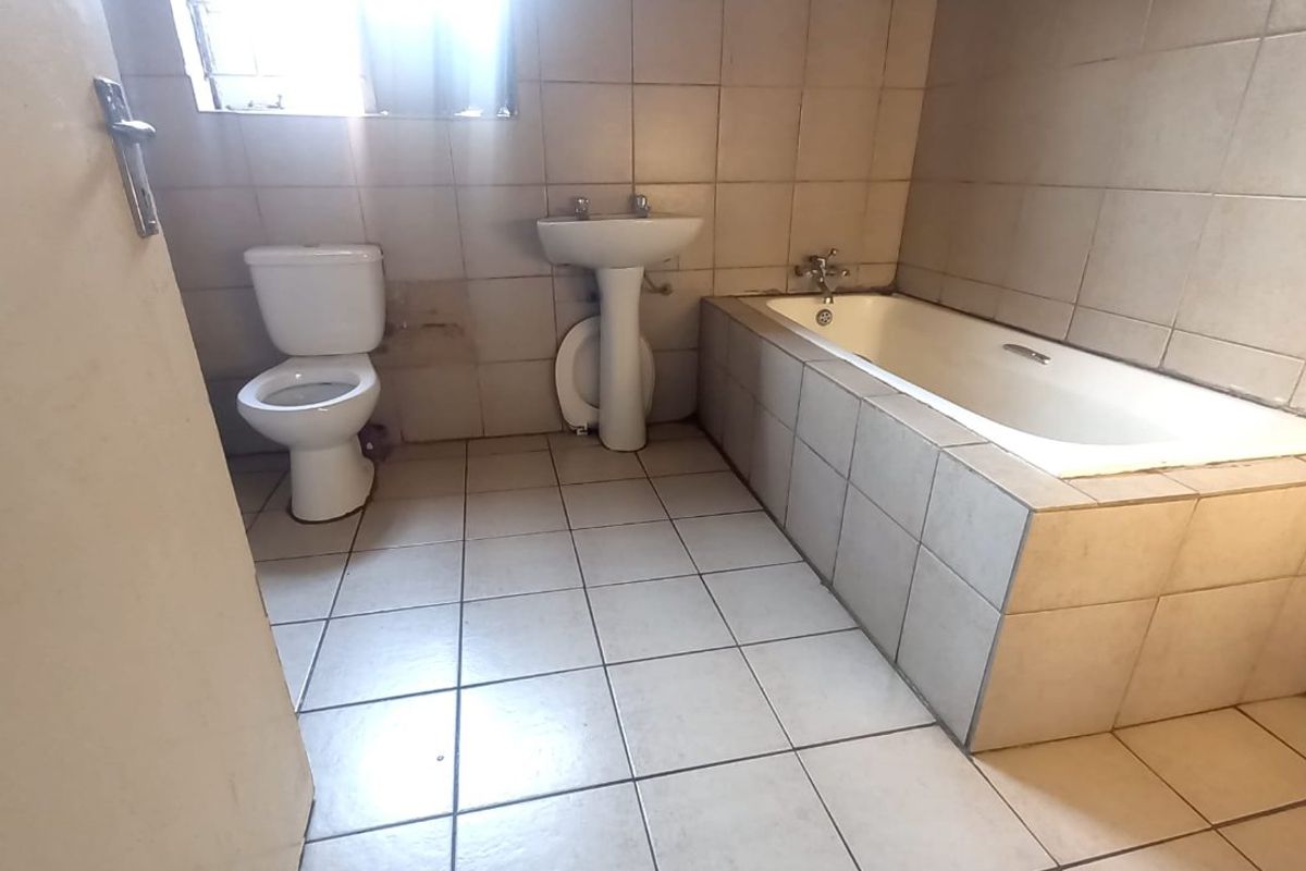 A functional bathroom featuring a built-in bathtub, pedestal basin, and toilet with neutral tiled finishes, requiring some cosmetic updates.
