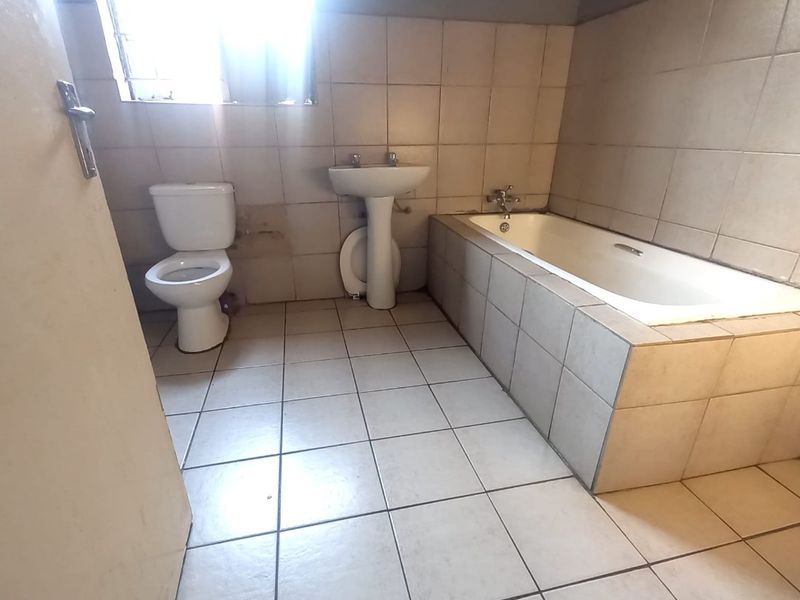 A functional bathroom featuring a built-in bathtub, pedestal basin, and toilet with neutral tiled finishes, requiring some cosmetic updates.