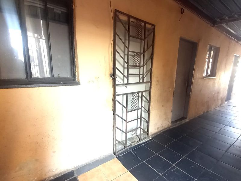 An exterior-facing hallway or walkway featuring tiled flooring, security gates, and metal window frames, showing signs of wear and requiring maintenance.