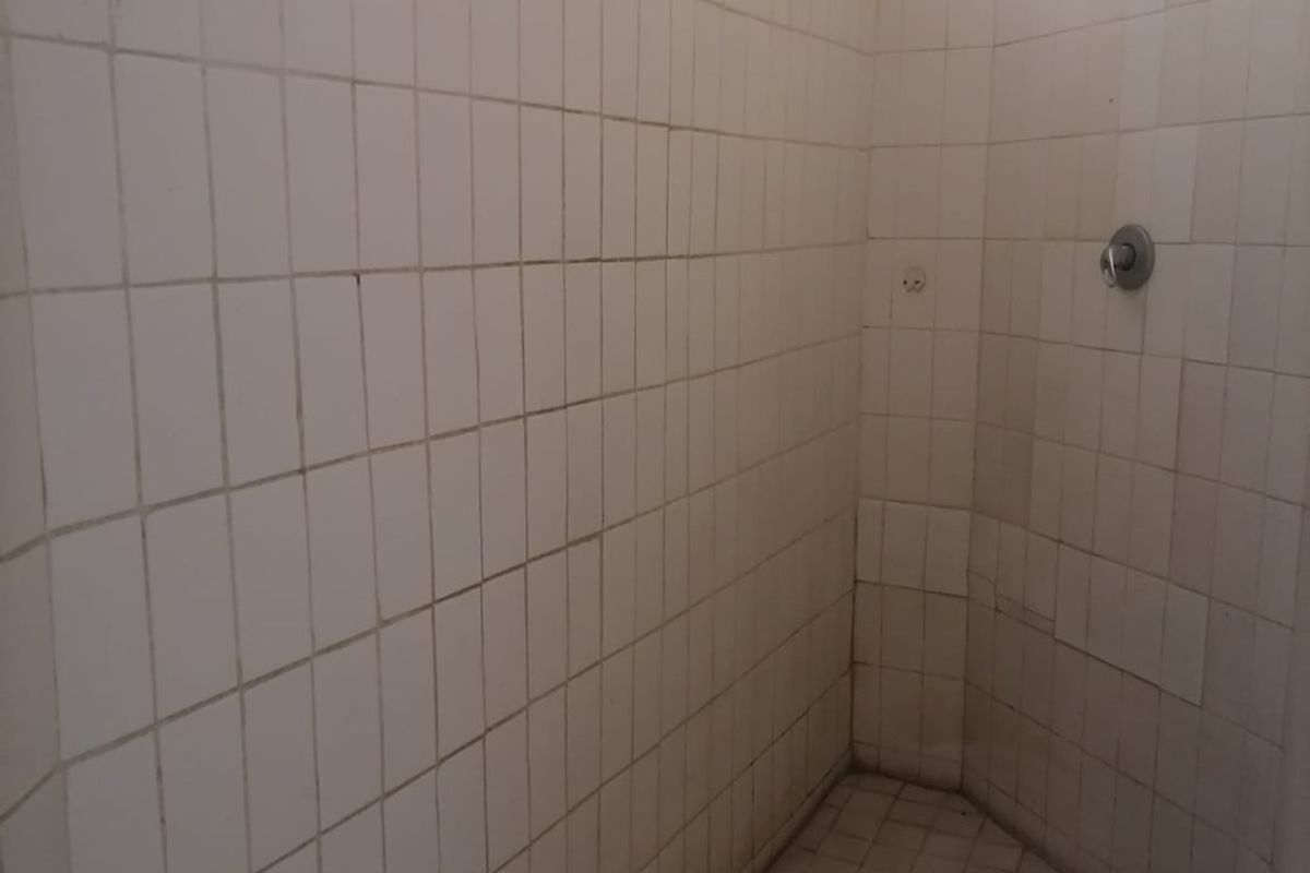 A basic walk-in shower area featuring white wall and floor tiles, a standard shower head, and mixer tap, requiring maintenance and cosmetic updates.
