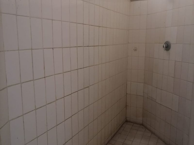 A basic walk-in shower area featuring white wall and floor tiles, a standard shower head, and mixer tap, requiring maintenance and cosmetic updates.