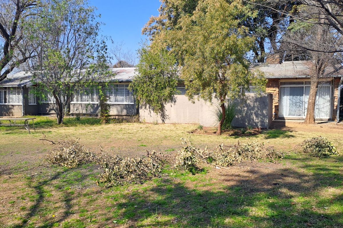 Rear view of a single-story house featuring stone masonry accents, steel-framed windows, and a large garden area requiring maintenance.