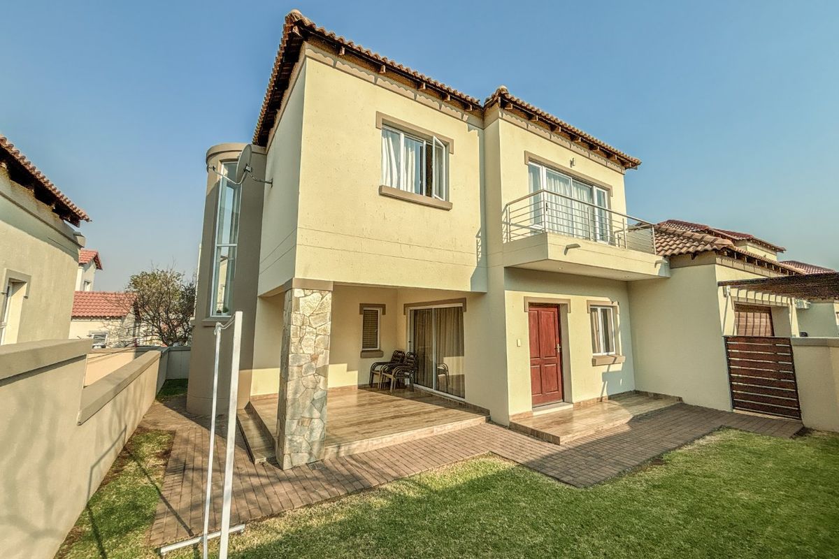 Rear view of a two-story residential house featuring a covered patio, a balcony with metal railings, stone-clad detailing, and a well-maintained garden area.