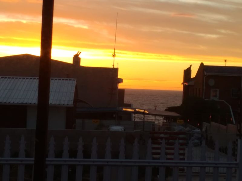 A scenic sunset view overlooking the ocean, framed by neighboring building silhouettes and a foreground fence.